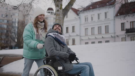 Glückliche-Kaukasische-Frau,-Die-Ihren-Behinderten-Freund-Im-Rollstuhl-Für-Einen-Spaziergang-In-Der-Winterstadt-Nimmt,-Während-Sie-Miteinander-Redet-Und-Lacht