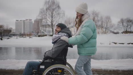 Side-view-of-happy-Caucasian-woman-taking-her-disabled-friend-in-wheelchair-for-a-walk-in-the-winter-city-and-talking-together