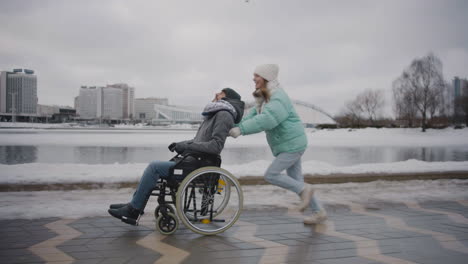 Glückliche-Frau,-Die-Mit-Ihrem-Behinderten-Freund-Im-Rollstuhl-Läuft-Und-Im-Winter-Gemeinsam-Spaß-In-Der-Stadt-Hat