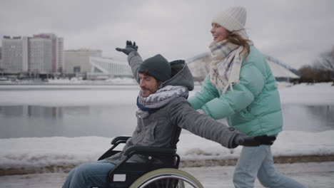 Glückliche-Frau,-Die-Mit-Ihrem-Behinderten-Freund-Im-Rollstuhl-Läuft-Und-Im-Winter-Gemeinsam-Spaß-In-Der-Stadt-Hat