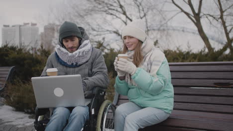 Disabled-man-in-wheelchair-watching-something-funny-on-laptop-computer-with-his-friend-at-urban-park-in-winter