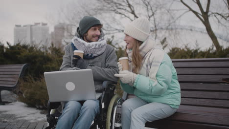 Behinderter-Mann-Im-Rollstuhl-Und-Sein-Freund-Beobachten-Etwas-Lustiges-Auf-Dem-Laptop,-Während-Sie-Im-Winter-Im-Stadtpark-Kaffee-Zum-Mitnehmen-Trinken