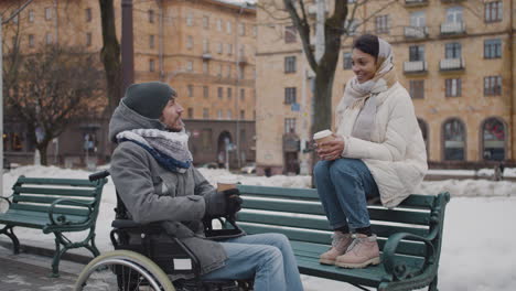 Muslim-woman-and-her-disabled-friend-in-wheelchair-drinking-takeaway-coffe-on-a-bench-in-city-in-winter