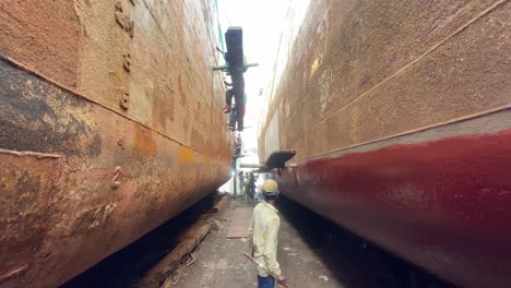 teenager-painting-ship-hull-painting-standing-between-two-rusty-old-ships-with-working-people-in-the-background-in-Dhaka,-Bangladesh