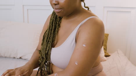 Close-up-view-of-African-american-woman-in-underwear-sitting-on-the-bed-spreading-moisturizing-cream-on-her-arms