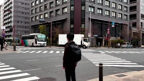 Young-Male-Asian-With-Mask-and-Backpack-Waiting-at-Zebra-Crossing,-Kyoto,-Japan