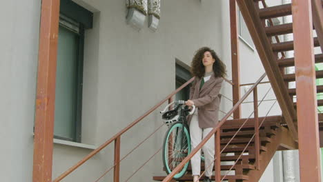 Curly-Caucasian-woman-in-formal-clothes-carrying-her-bike-down-the-stairs