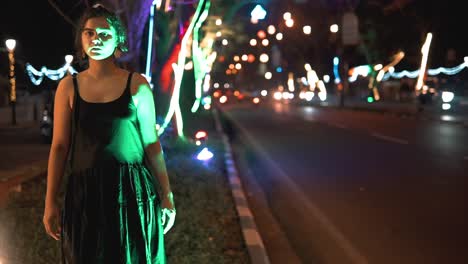 Brunette-woman-in-a-black-dress-walking-towards-the-camera-in-a-street-scene-full-of-cars-and-colored-lights