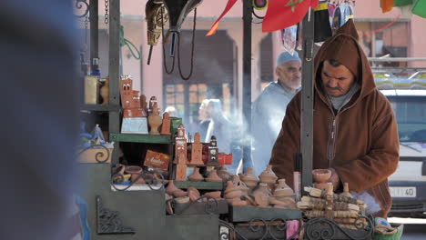 Vendor-with-brown-hood-at-stall-with-incense-smoke-blowing-around,-Marrakesh