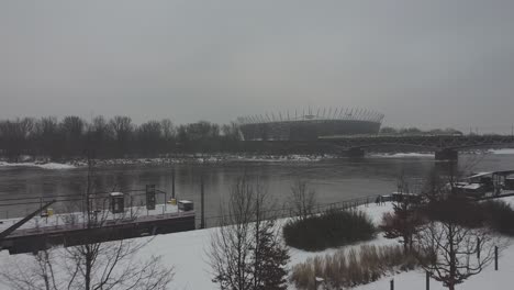 Drohnenvideo-Des-Warschauer-Nationalstadions,-Während-An-Einem-Verschneiten-Tag-über-Der-Weichsel-Ein-Zug-Auf-Einer-Brücke-Vorbeifährt1