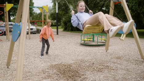 Little-girl-with-down-syndrome-wearing-hoodie-swinging-on-a-swing-in-the-park-on-a-windy-day.-Her-male-friend-pushing-her