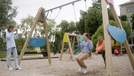 Little-girl-with-down-syndrome-wearing-hoodie-swinging-on-a-swing-in-the-park-on-a-windy-day.-Her-male-friend-pushing-her-while-her-female-friend-take-a-photo