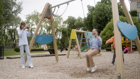 Little-girl-with-down-syndrome-wearing-hoodie-swinging-on-a-swing-in-the-park-on-a-windy-day.-Her-male-friend-pushing-her-while-her-female-friend-take-a-photo