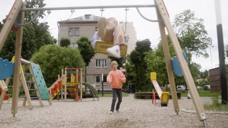 Front-view-of-little-girl-with-down-syndrome-wearing-hoodie-swinging-on-a-swing-in-the-park-on-a-windy-day.-Her-male-friend-pushing-her-while-her-female-friend-take-a-photo