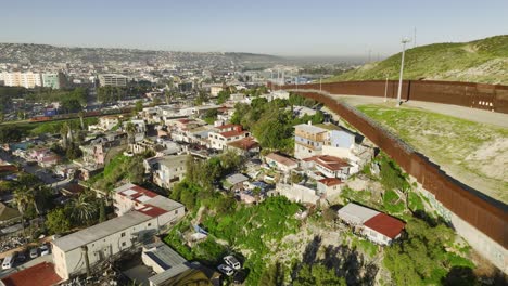 Aerial-view-following-the-wall,-over-a-poor-area,-towards-the-border-station-in-sunny-San-Ysidro,-Tijuana,-Mexico