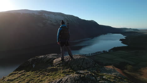 Hiker-on-rocky-outcrop-with-reveal-of-dark-lake-and-green-valley-plus-snowy-mountains-and-natural-lens-flare-at-Wasdale-Lake-District-UK