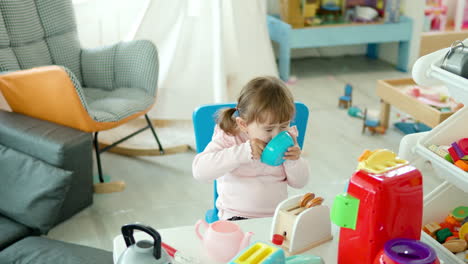Playful-Little-Girl-Pretends-Eating-From-Toy-Plate-with-Spoon-Sitting-on-Chair-by-the-Table-with-Kitchenware-Cooking-Toys