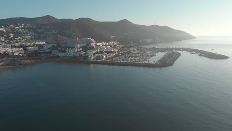 drone-captures-stunning-footage-of-a-coastal-town-port,-with-magnificent-mountain-range-and-charming-housing-estates-nestled-in-background-during-early-morning