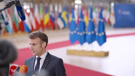 French-president-Emmanuel-Macron-talking-to-the-press-with-in-the-background-the-European-flags-and-EU-member-state-flags-at-the-European-Council-summit-in-Brussels,-Belgium