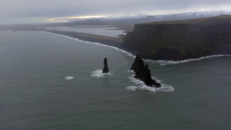 Aerial:-Slow-panning-shot-of-Reynisfjara-black-sand-beach-and-Reynisdrangar-sea-stacks-in-Iceland