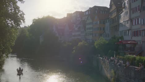 Boat-Passing-by-as-are-People-Relaxing-On-Wall-In-Tubingen,-Germany-in-4K-Downtown-Home-of-Europes-Oldest-University-At-Sunset
