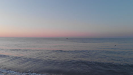 Blauer-Horizont-Sonnenuntergangsdrohne-Mit-Ruhigen-Wellen,-Skyline-Ufer-Ins-Meer