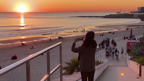 view-of-woman-recording-huge-sunset-with-unrecognised-people-in-background-at-Carcavelos-beach,-small-waves-form-and-break,-beautiful-Atlantic-ocean,-deep-yellow-sunset-on-water-reflections,-Portugal