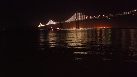 Gimbal-tilting-up-shot-of-a-ferry-under-the-Bay-Bridge-at-night-in-San-Francisco,-California