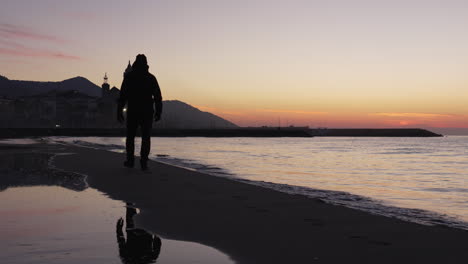 man-walks-along-tranquil-sea,-dusky-coastline-stretches-out-before-him,-leading-towards-silhouette-of-a-church-set-against-the-majestic-mountain-range