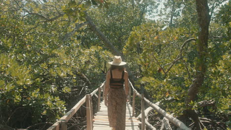 Slow-motion-from-behind-of-an-attractive-Caucasian-woman-in-a-straw-hat-crossing-a-wooden-bridge-in-a-green-forest