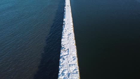 Revealing-aerial-view-of-snow-and-ice-covered-concrete-pier-in-the-calm-Baltic-sea,-Port-of-Liepaja-on-a-sunny-winter-day,-slow-wide-angle-drone-shot-moving-forward-camera-tilt-up