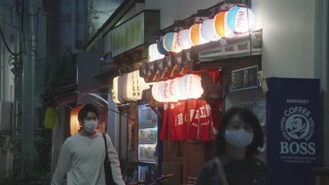 Man-And-Woman-Wearing-Face-Mask-Walking-And-Passing-By-Japanese-Izakaya-With-Illuminated-Paper-Lanterns-At-Night-During-Pandemic-In-Tokyo,-Japan