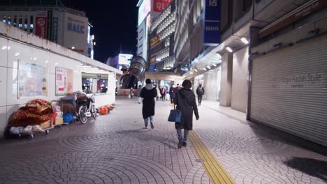 Wide-angle-shot-behind-a-woman-walking,-and-other-people-wearing-masks,-outside-a-quiet-Shinjuku-Station-at-night-passing-a-homeless-person-sleeping-outside-and-shops-closed