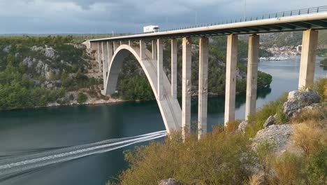 A-powerboat-is-passing-underneath-Krka-bridge-near-Skradin-town,-Croatia-while-truck-is-driving-above-on-the-highway