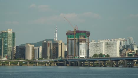 Hangang-Flussufer-Mit-Autoverkehr-Auf-Der-Gangbyeon-Schnellstraße,-Mapodaegyo-Brücke-Und-Mapo-Gu-Stadtsilhouette-Mit-Wahrzeichen-Seoul-Namsan-Tower-über-Blauem-Himmel,-Südkorea