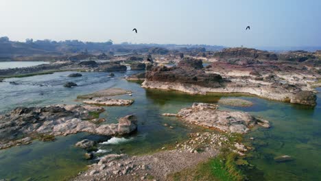 Drone-backward-moving-shot-over-beautiful-Narmada-river-in-Gujrat,-India-passing-through-rock-structures-on-a-bright-sunny-day