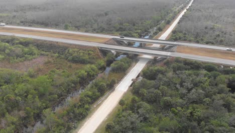 highway-bridge-over-canal-dirt-road-overpass-everglades-expressway-florida-aerial-drone-tilt