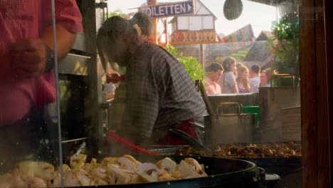 Two-cooks-making-food-behind-glass-at-outdoor-street-festival-in-downtown-Stuttgart-at-sunset,-Germany,-Europe,-panning-view-angle