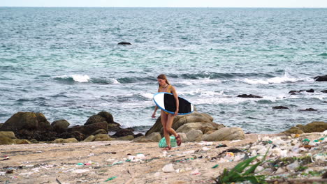 Young-surfer-in-bikini-walk-in-sandal-on-beach-with-trash-plastic-waste-and-ocean-pollution,-female-walking-hold-surf-board