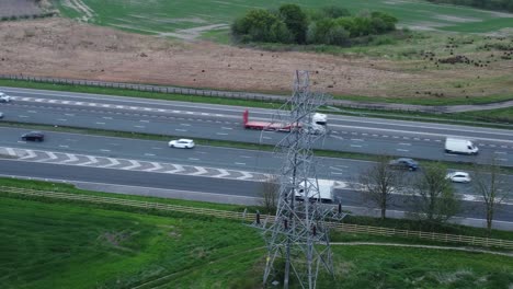 Vehicles-on-M62-motorway-passing-pylon-tower-on-countryside-farmland-fields-aerial-orbit-right-view