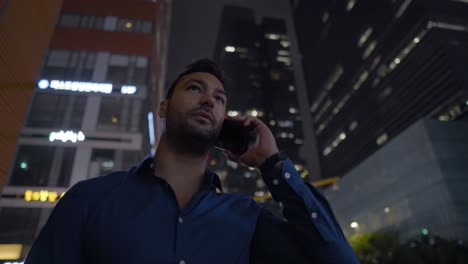 Young-businessman-is-having-a-phone-talk-while-standing-by-high-rise-skyscrapers-in-city-downtown-at-night