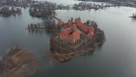 AERIAL:-Rotating-Shot-of-Red-Colour-Trakai-Island-Castle-Surrounded-with-Lake-and-Houses-in-the-Distance