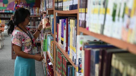 Eine-Asiatische-Frau-Nähert-Sich-Den-Bücherregalen,-Um-Sich-Ein-Buch-Zu-Schnappen,-Während-Sie-Kichert-Und-Ihr-Lieblingsbuch-Findet