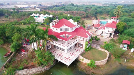 Close-up-of-red-tiled-roof-and-white-luxurious-vacation-home-surrounded-by-forest-and-native-vegetation-with-beautiful-lake-and-pool-in-Vadodara,-India