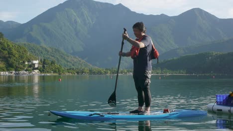 Asian-man-on-stand-up-paddle-board-rowing-across-lake-surrounded-by-mountains