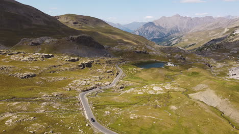 Slowly-descending-drone-on-the-top-of-the-alps,-a-road-leads-to-a-deep-blue-mountain-lake