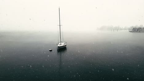 Surreal-scene-of-snow-falling-over-Maggiore-lake-and-moored-sailing-boat-and-ferryboats,-Italy