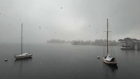 Snow-falling-over-Maggiore-lake-and-anchored-sailing-boat-and-ferry,-Italy
