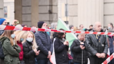 Red-And-White-Barricade-Tape-With-Protesters-In-Background-At-Piazza-XXV-Aprile-Amidst-Pandemic-In-Milan,-Italy