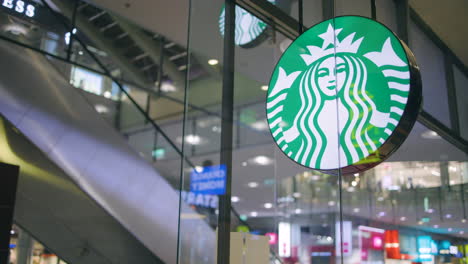 Slomo-of-Starbucks-logo-on-sign-by-glass-wall-and-escalator-in-mall